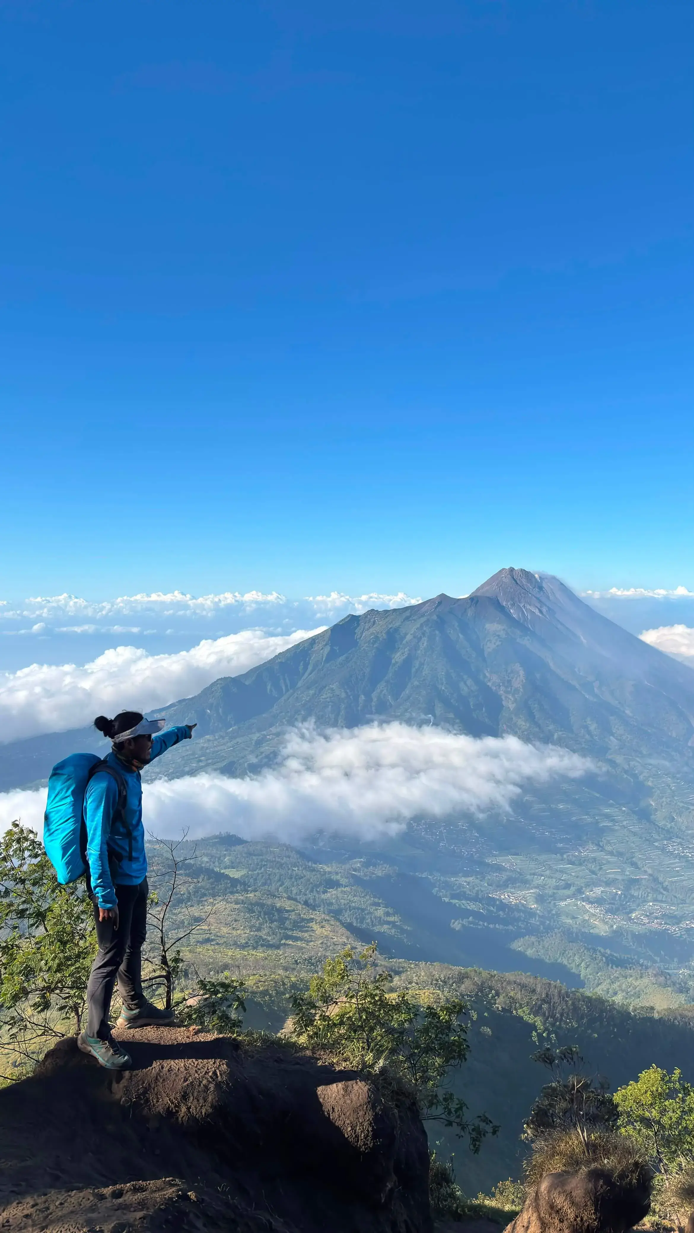 A hiker standing on a ridge and pointing towards the majestic peak of Mount Merapi during a Mount Merbabu trekking tour.